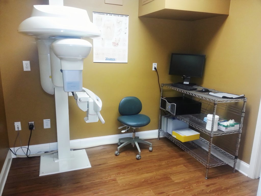 Clinical room housing a white panoramic X-ray machine and a mobile workstation on hardwood flooring
