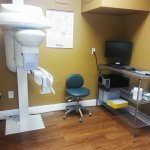 Clinical room housing a white panoramic X-ray machine and a mobile workstation on hardwood flooring